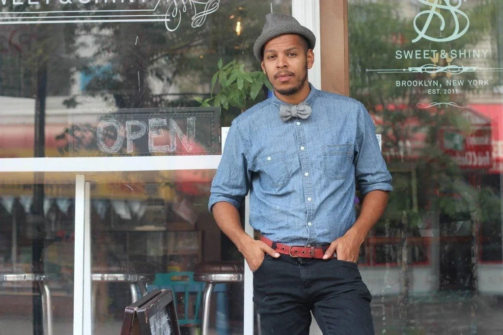 Hombre con pajarita gris, camisa azul de manga larga y sombrero gris posando frente a una tienda con cartel abierto