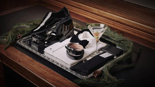 Conjunto elegante para hombre con zapatos negros, camisa blanca, pajarita negra, cinturón, gemelos, calcetines y copa de martini sobre bandeja con decoración navideña