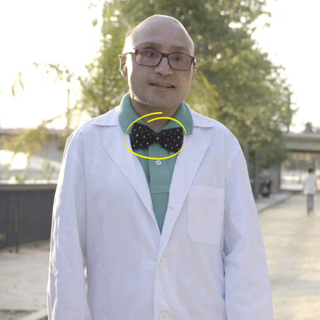 Hombre con bata blanca y pajarita negra con estampado colorido, camisa verde, al aire libre en parque
